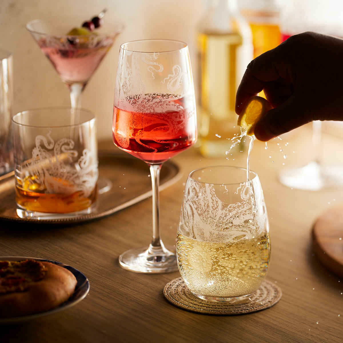 Lucy the Octopus stemless wine, stemmed red wine, and rocks glasses in etched crystal from Caskata with a hand squeezing a lemon into the glass.