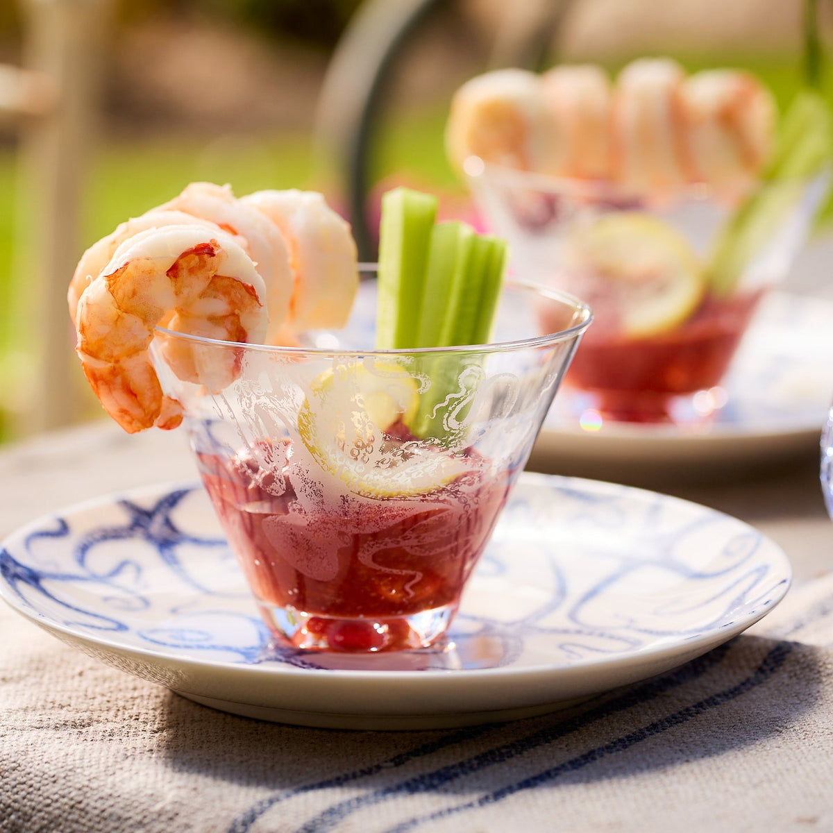 A Caskata Artisanal Home Lucy Stemless Martini Glass containing cocktail sauce garnished with shrimp, celery sticks, and lemon slices, placed on a decorative plate with a blue pattern. Another similar glass is partially visible in the background.