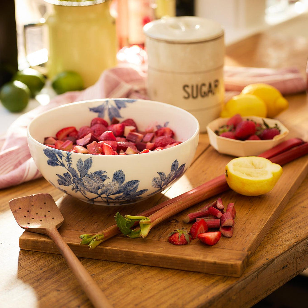 A premium Caskata Artisanal Home Arbor Vegetable Serving Bowl of chopped strawberries sits on a wooden cutting board with a spoon, fresh rhubarb, lemon halves, and a sugar container in a kitchen setting.