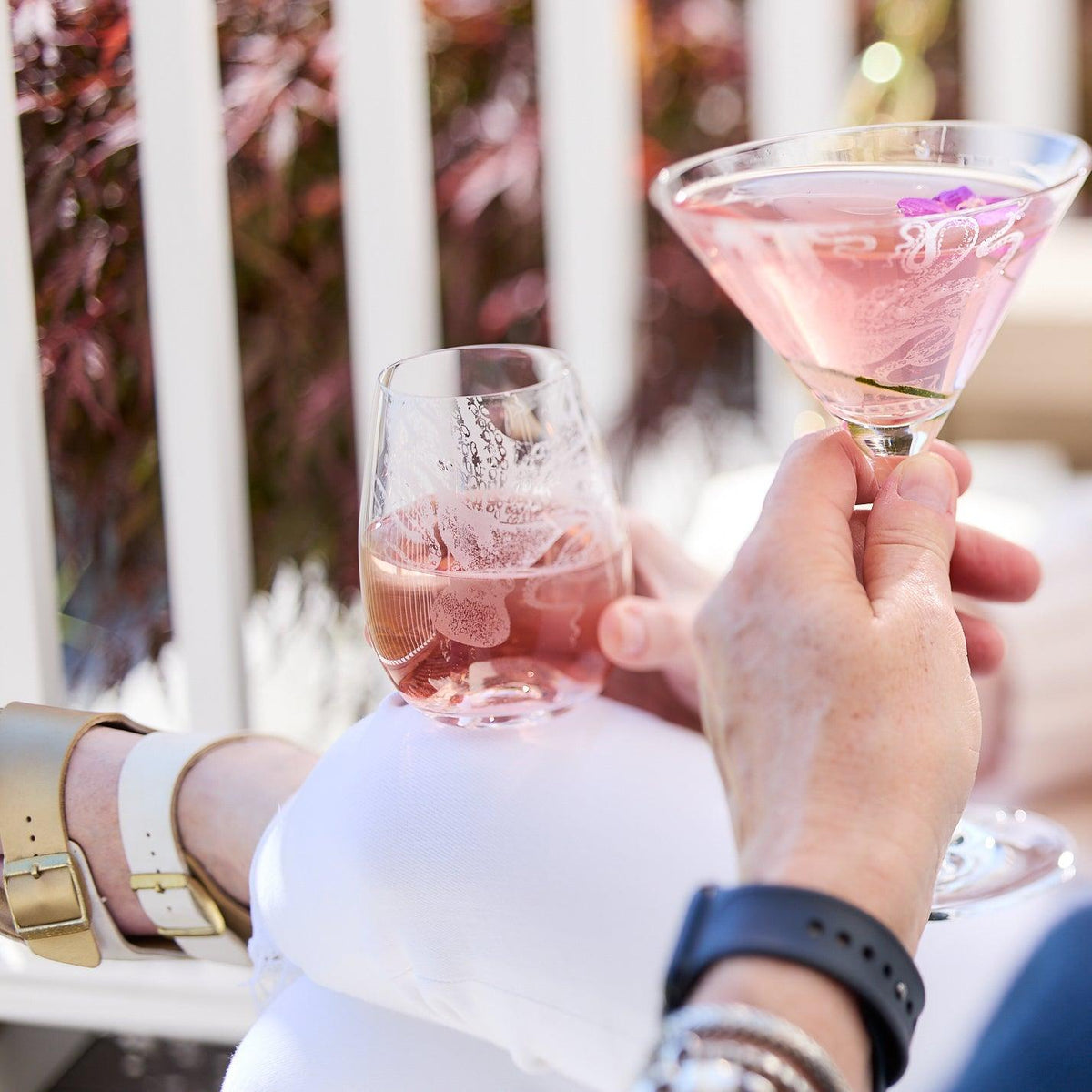 Two people holding drinks, one in a Lucy Stemless Wine Glass by Caskata Artisanal Home and the other in a martini glass, while sitting outdoors near a railing with foliage in the background. One person is wearing a smartwatch and bracelets.