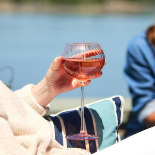 A person holds a Quinn Rose Red Wine Glass from Caskata Artisanal Home filled with pink liquid, likely rosé, against a blurred outdoor backdrop. The lead-free crystal of the glass adds an extra touch of elegance to the scene.