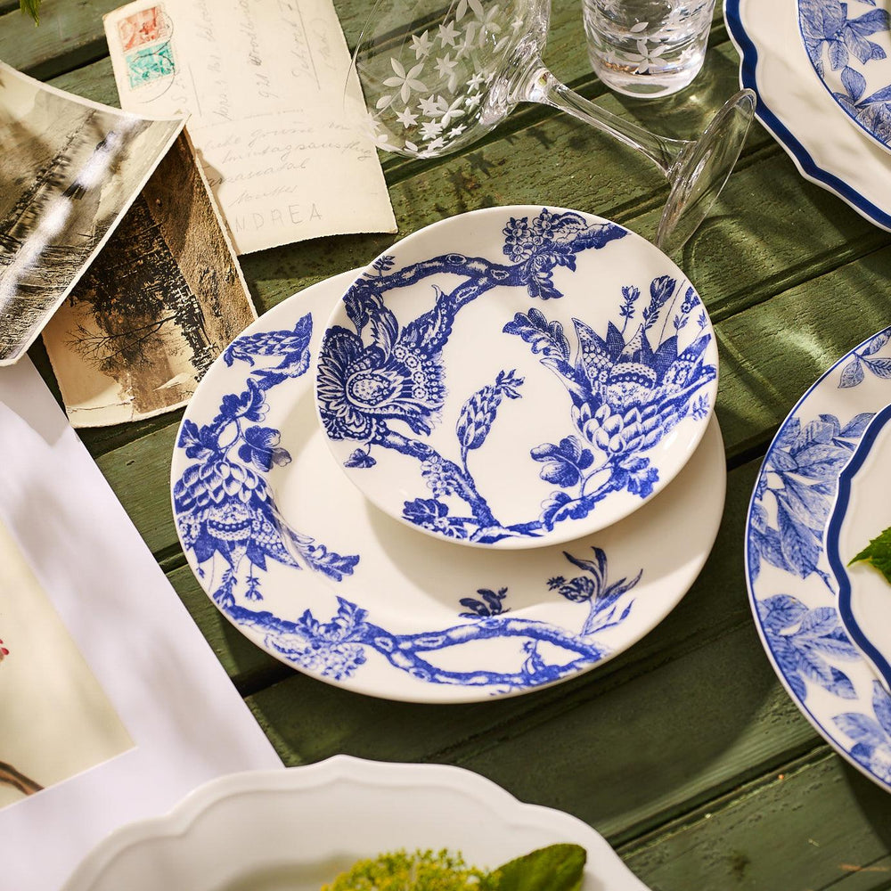 A set of Arcadia Rimmed Salad Plates by Caskata Artisanal Home, crafted from premium porcelain and featuring intricate floral and bird designs in blue-and-white, sits on a wooden table surrounded by letters and a glass of water. This elegant collection brings an heirloom feel to any setting.