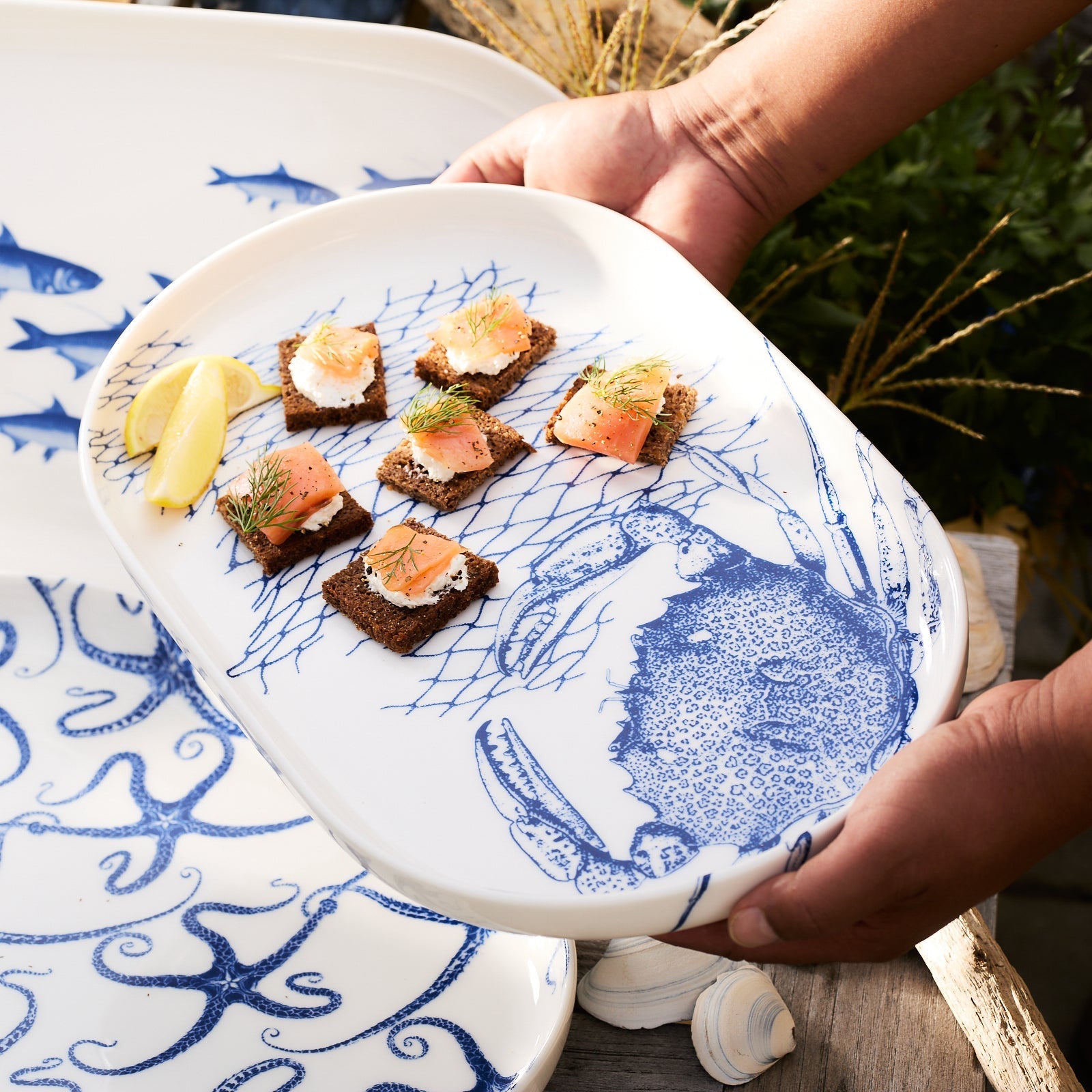 The Caskata Crab Crudité Set features a white porcelain platter and small bowl with blue crab and net illustrations—perfect coastal dinnerware or a charming addition to seaside decor.