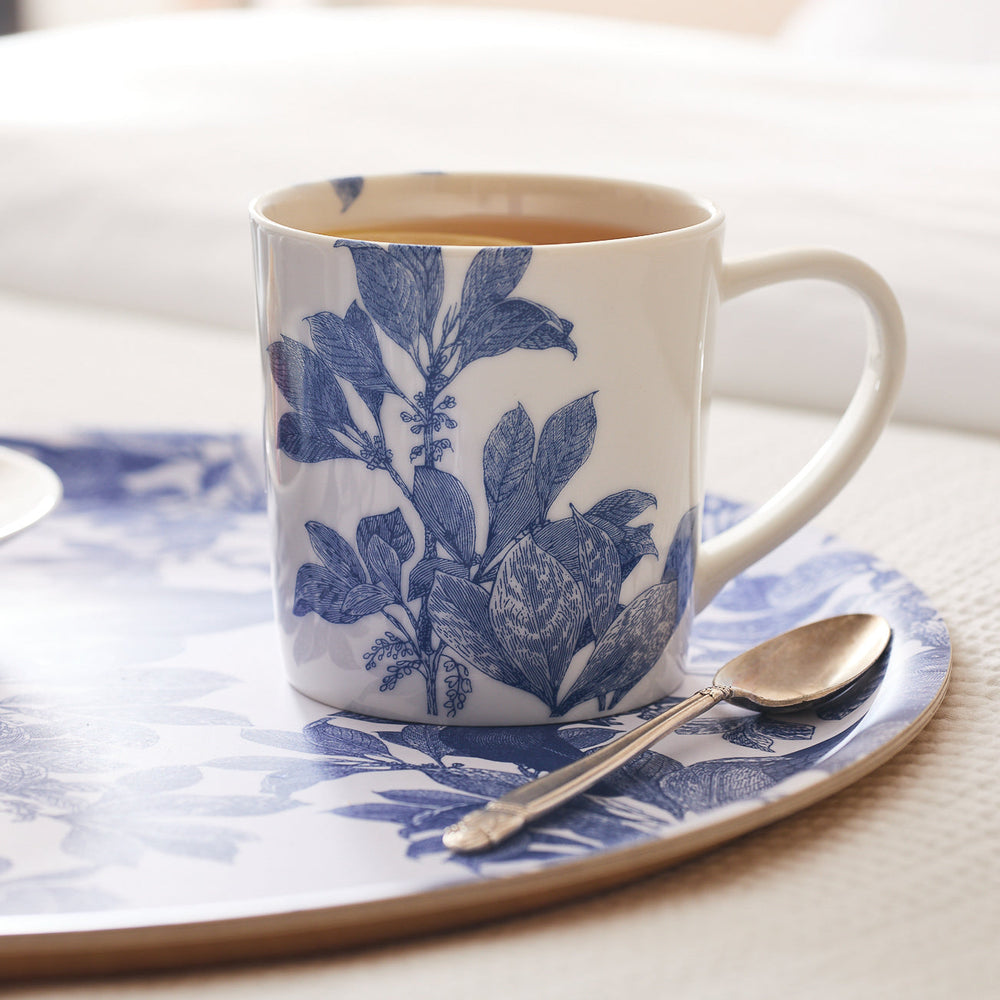 A generously sized Arbor Mug from Caskata Artisanal Home, featuring a blue floral pattern and crafted from premium porcelain, holds tea and is placed on a matching Arbor tray with a silver spoon beside it.