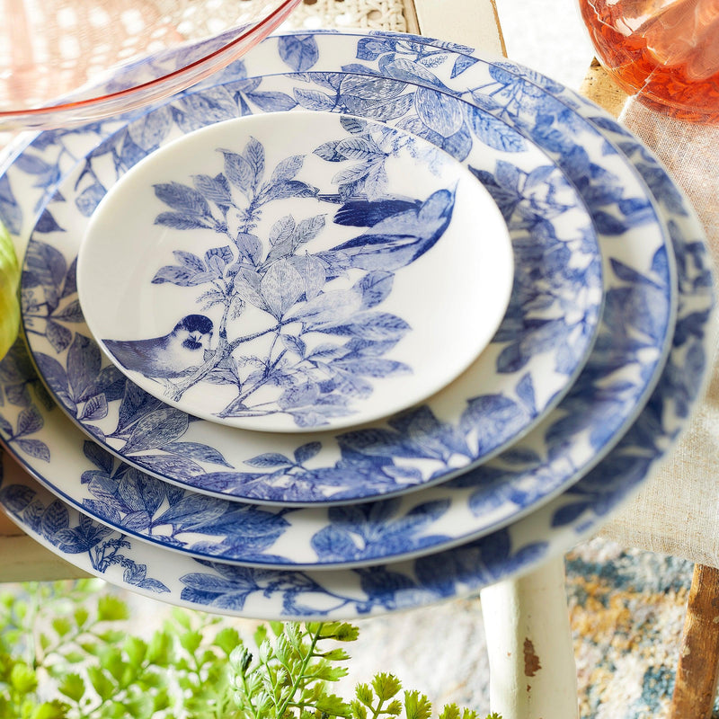 Close-up of a stack of Caskata Artisanal Home Arbor Rimmed Dinner Plates with blue floral and bird patterns, arranged on a table with a light-colored linen cloth underneath. A green plant is partially visible in the foreground, highlighting the heirloom-quality dinnerware's delicate botanical details.