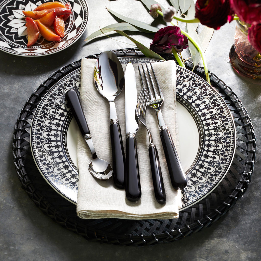 The Casablanca Table for 8 by Caskata features a black and white patterned porcelain dinnerware set with a woven charger, napkin, and black-handled cutlery. Dishwasher and microwave safe. Flowers and a bowl of sliced strawberries complete the look.