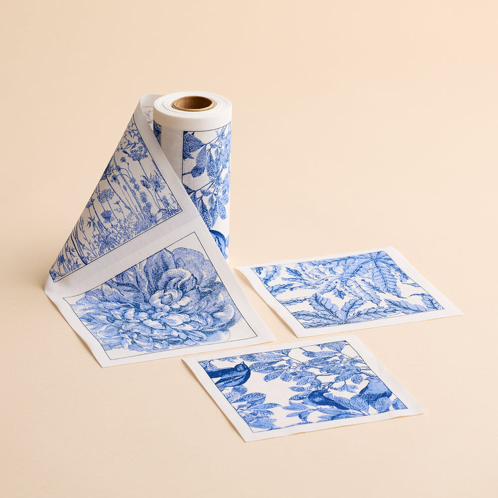 A roll of MY DRAP’s Botanicals Cocktail Napkin Roll is unspooled, with three individual square napkins featuring blue and white floral and botanical designs placed beside it on a beige surface—evoking the charming motifs often found on handcrafted cotton napkins.