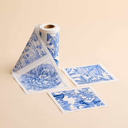 A roll of MY DRAP’s Botanicals Cocktail Napkin Roll is unspooled, with three individual square napkins featuring blue and white floral and botanical designs placed beside it on a beige surface—evoking the charming motifs often found on handcrafted cotton napkins.