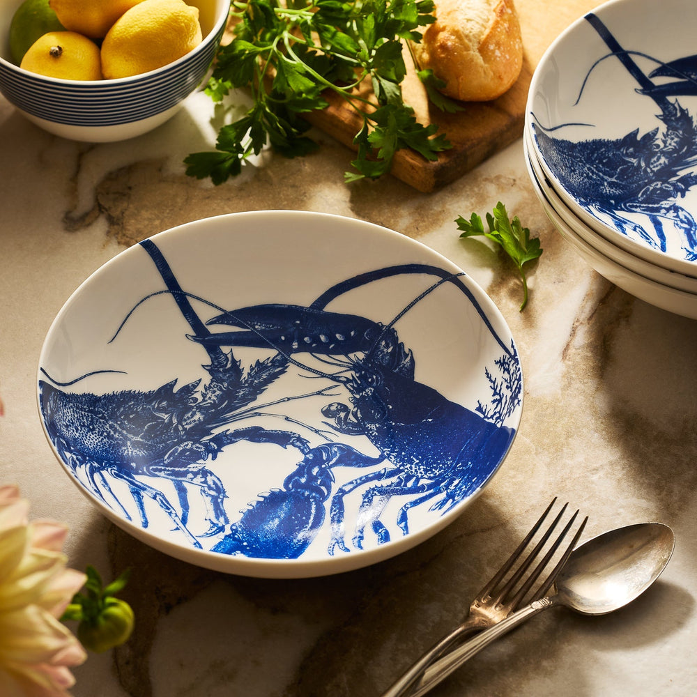 The Caskata Lobster Entrée Bowl, showcasing a blue lobster design, rests on marble with parsley, a baguette, lemons, a spoon, and a fork. This charming, dishwasher-safe porcelain bowl enhances any tablescape.