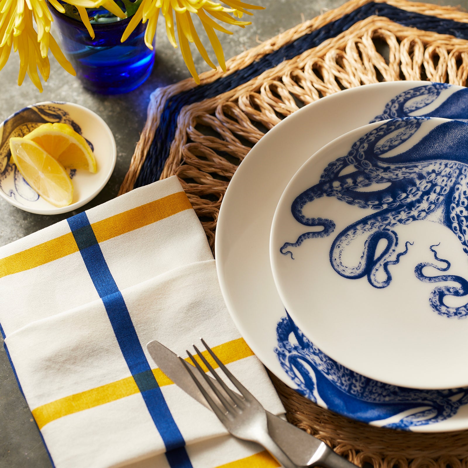 The Lucy Table for 12 by Caskata features octopus pattern dinnerware, a woven placemat, yellow and blue napkins, cutlery, a lemon slice, and yellow flowers in a blue vase.