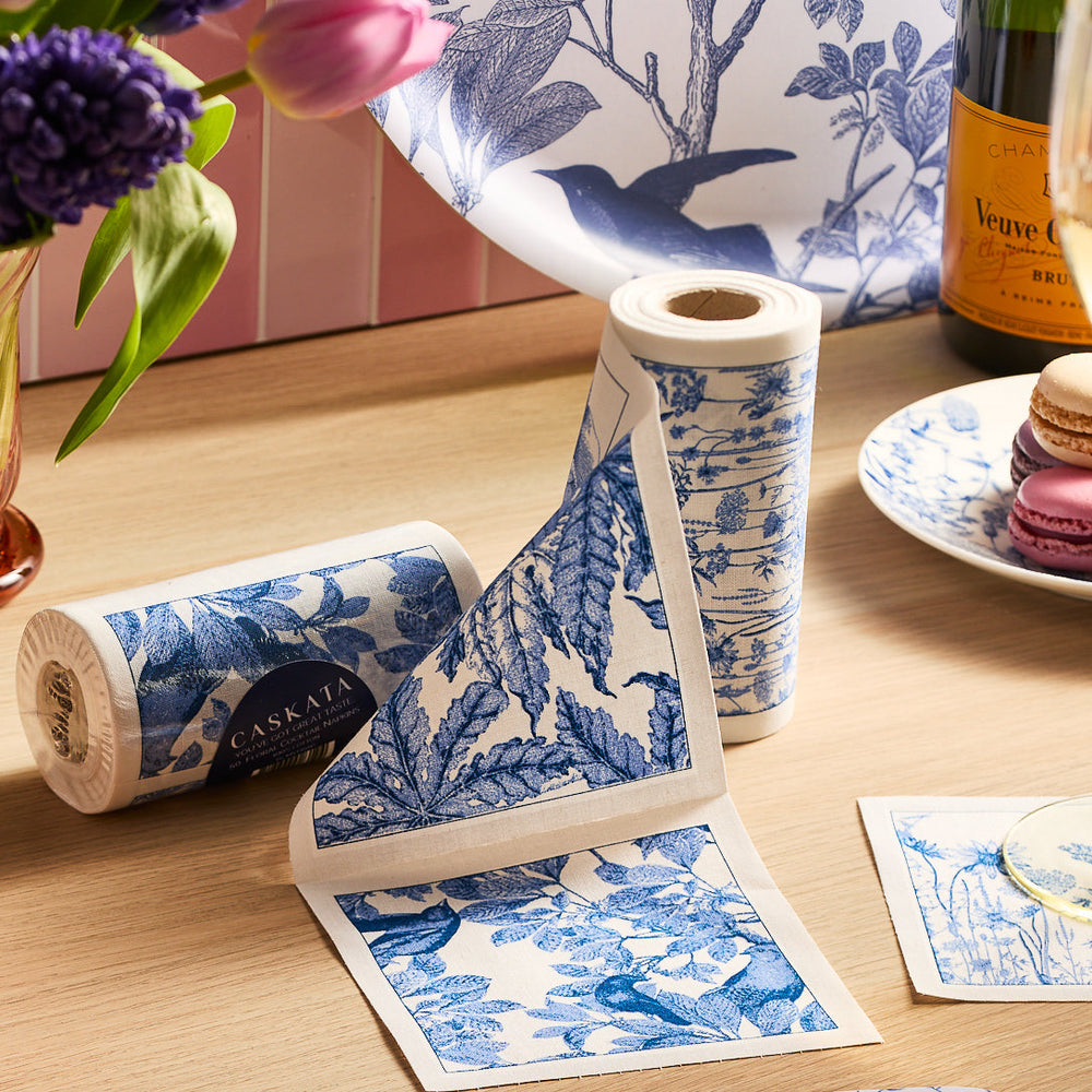 MY DRAP's Botanicals Cocktail Napkin Roll featuring floral and bird patterns in blue and white adorns the table, complemented by reusable napkins, vibrant flowers, macarons, and a bottle of champagne ready for celebration.