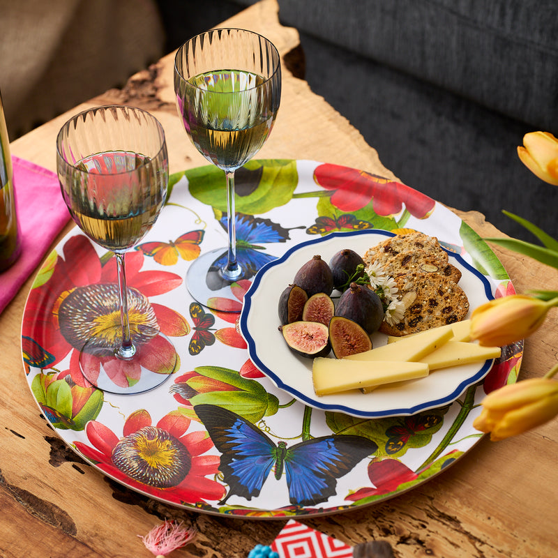 Two wine glasses rest on the Caskata Butterflies Large Round Birchwood Tray with figs, cheese slices, and crackers on a plate. Yellow tulips are partially visible in the foreground.
