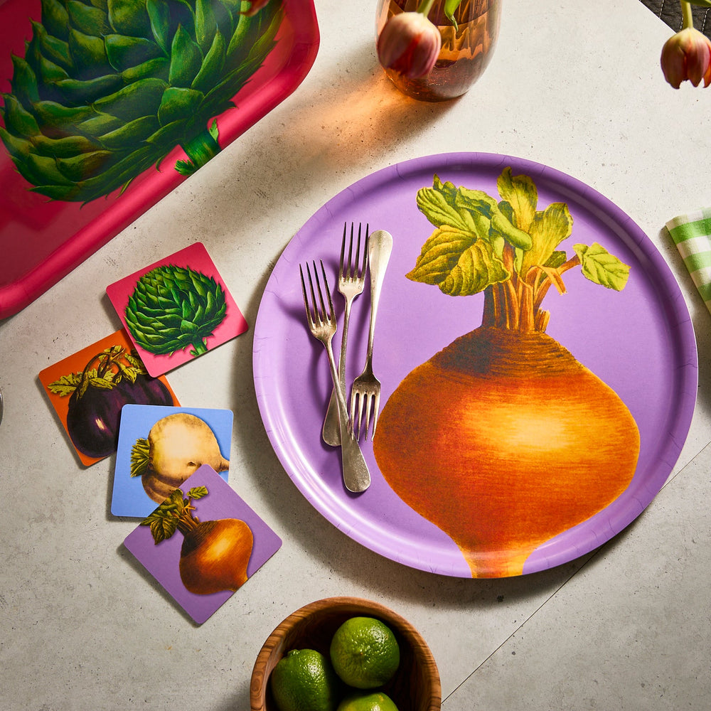 A vibrant tabletop display features a large plate with a turnip design, surrounded by forks and artichoke coasters. Limes rest in a wooden bowl, and the scene is enhanced by Caskata's Golden Beet Large Round Birchwood Tray, celebrating sustainable style and fresh veggies.