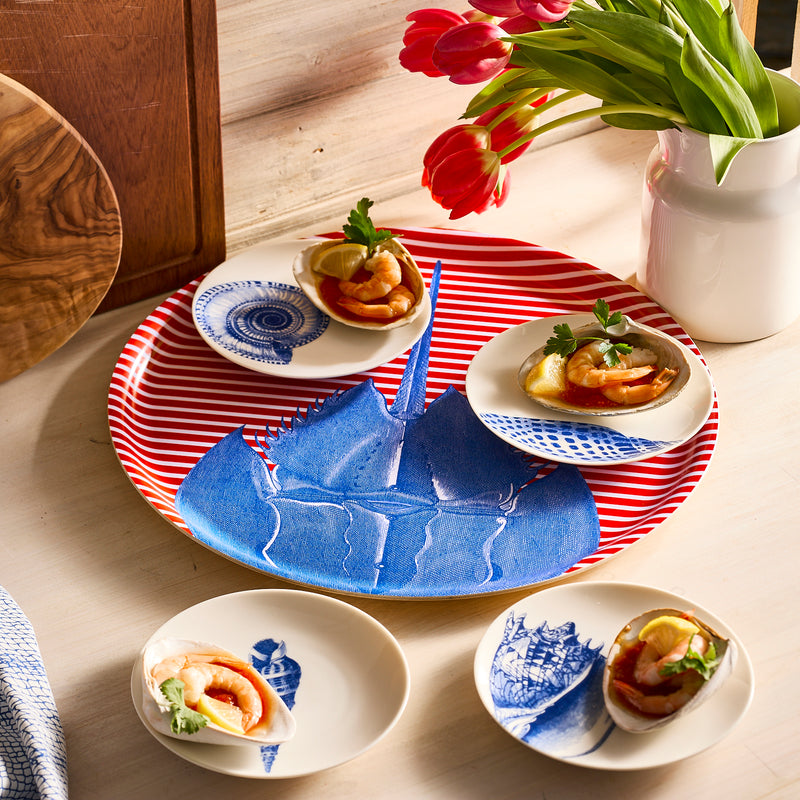 Shells Small Plates by Caskata Artisanal Home, adorned with seafood appetizers and garnishes, rest on the wooden table. Red tulips in a white vase enhance the striped tray with a blue design, adding elegance to this heirloom-quality dinnerware setting.
