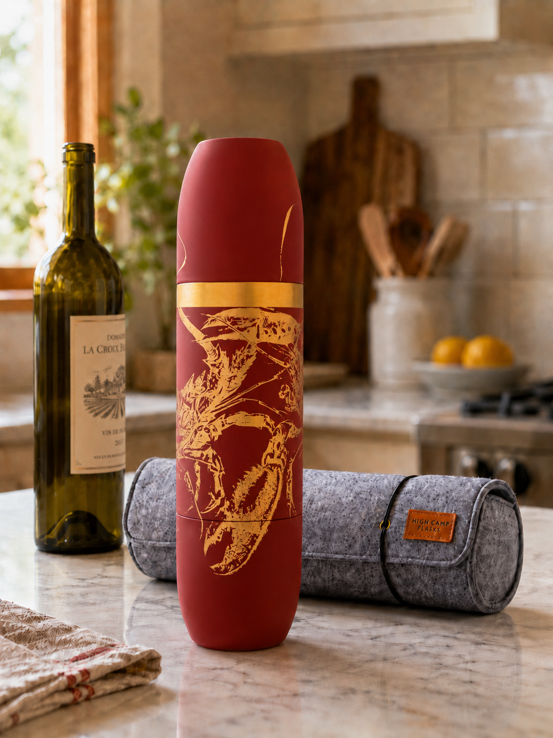 Caskata X High Camp red Lobster Wine flask engraved in gold on a kitchen counter with a bottle of wine and a wool carrying case.