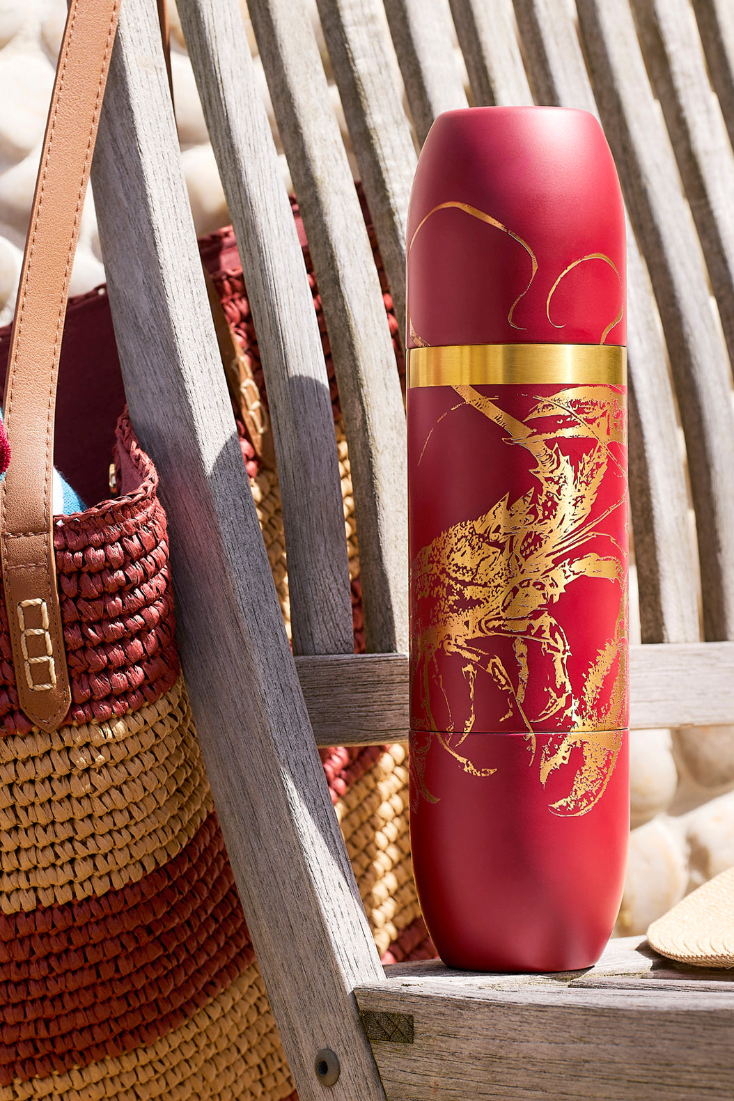 Caskata x High Camp wine flask in Rosso (red) with two lobsters engraved in gold, on a weathered wooden chair with a straw bag