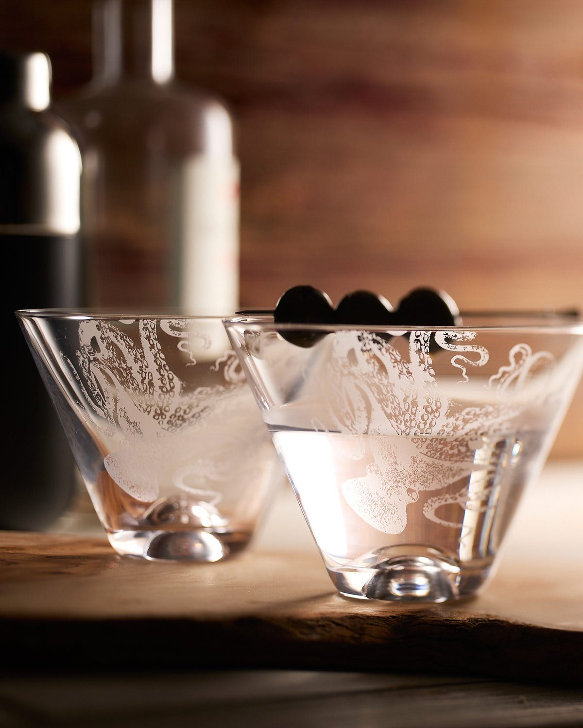 Caskata's Lucy the Octopus Crystal Etched Stemless martini glasses sold as a set of two on a bar with a cocktail in the foreground softly lit.