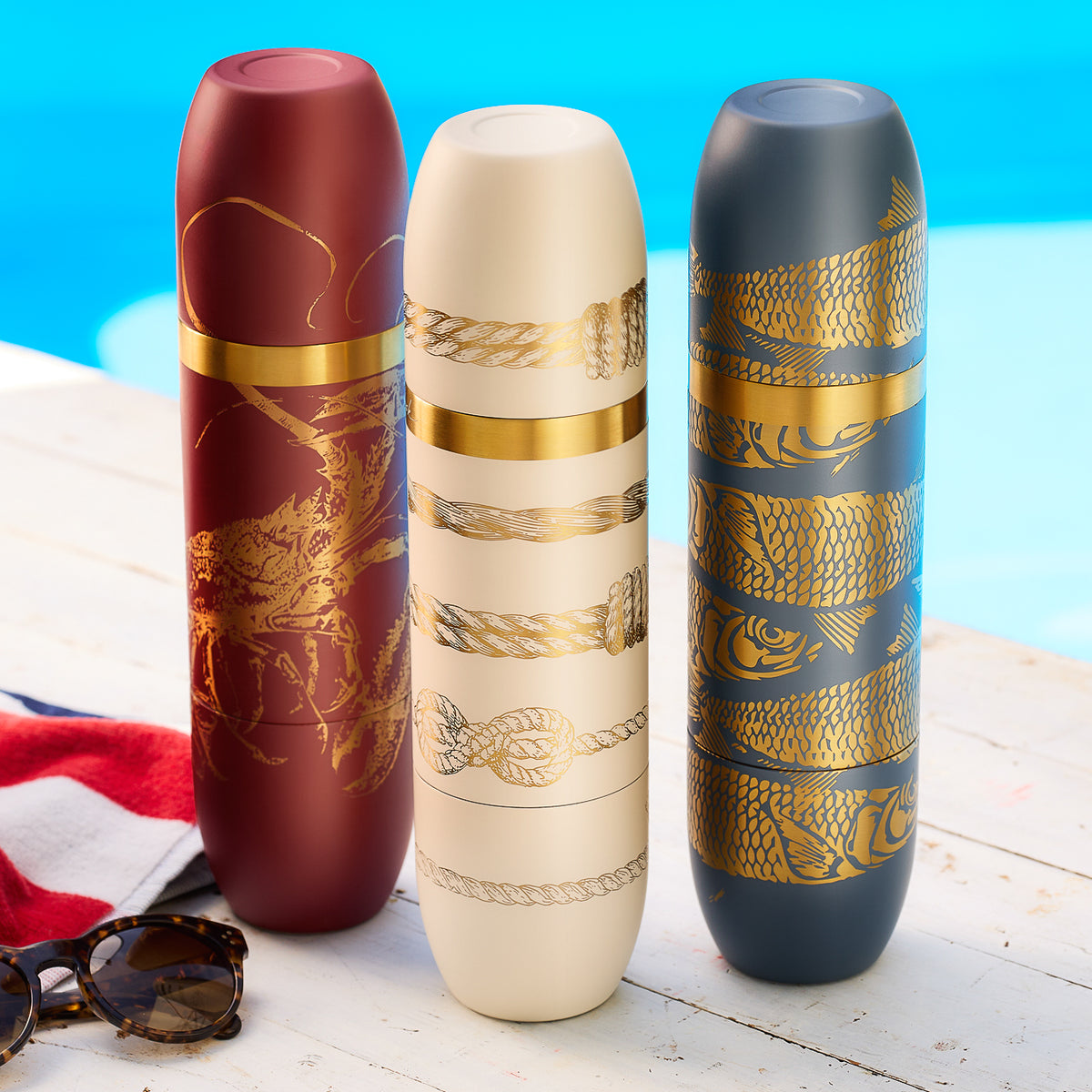 Three Caskata x High Camp Wine Flasks, Including Crema Sailors Knots, Sardines, and Lobsters, all engraved with gold nautical designs sit on a white wooden surface by the pool, alongside sunglasses and a red, white, and blue striped towel.