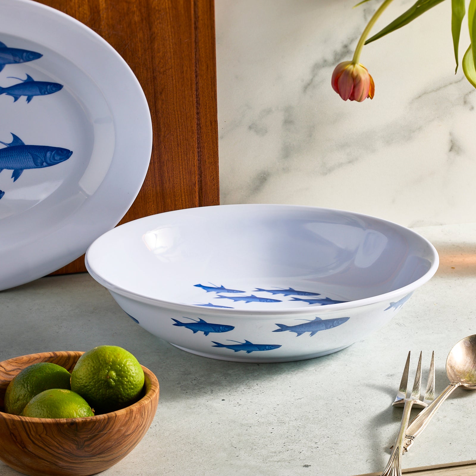 The School of Fish Entertaining Melamine Bundle by Caskata rests on a countertop, complemented by limes in a wooden bowl, cutlery, a flower, and a matching plate in the background.