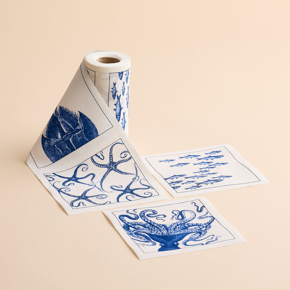 The MY DRAP Coastal Cocktail Napkin Roll features charming blue sea-themed designs, including fish, a starfish, and coral, unrolled on a beige surface—perfect for those seeking eco-friendly napkins with coastal patterns.