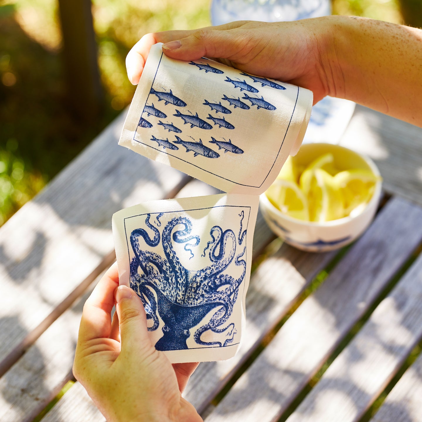 Two hands hold Caskata’s “Everything’s Going Swimmingly” Bundle ceramic cups, one with a School of Fish pattern and the other with an octopus, above a wooden outdoor table with a bowl of lemon slices.