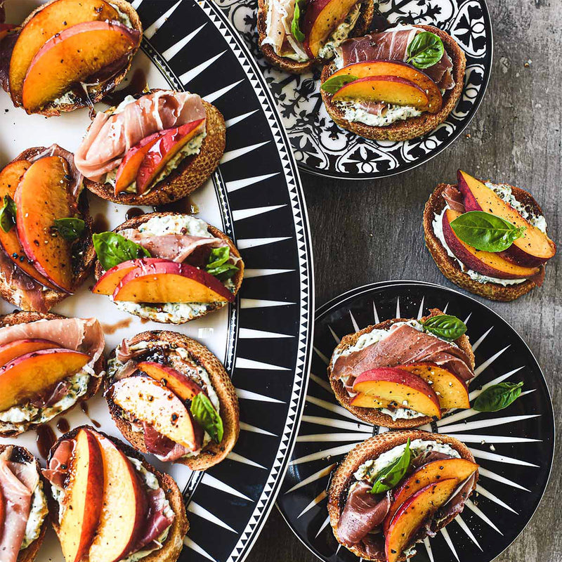 Small plates of open-faced sandwiches topped with peach slices, prosciutto, ricotta cheese, and basil are arranged on a table set with Caskata Artisanal Home Marrakech Small Plates.