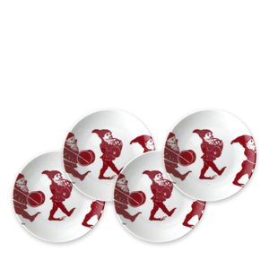 Four white plates, part of an heirloom-quality dinnerware holiday collection, each feature an illustration of a red gnome holding a drum. They are called Elves Small Plates by Caskata Artisanal Home.