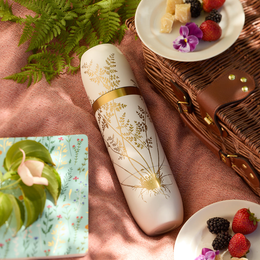 The Caskata x High Camp Crema Fairy Fern Wine Flask by Caskata rests on a picnic blanket beside a wicker basket, fresh berries, a decorative plate, and fern leaves in bright daylight.