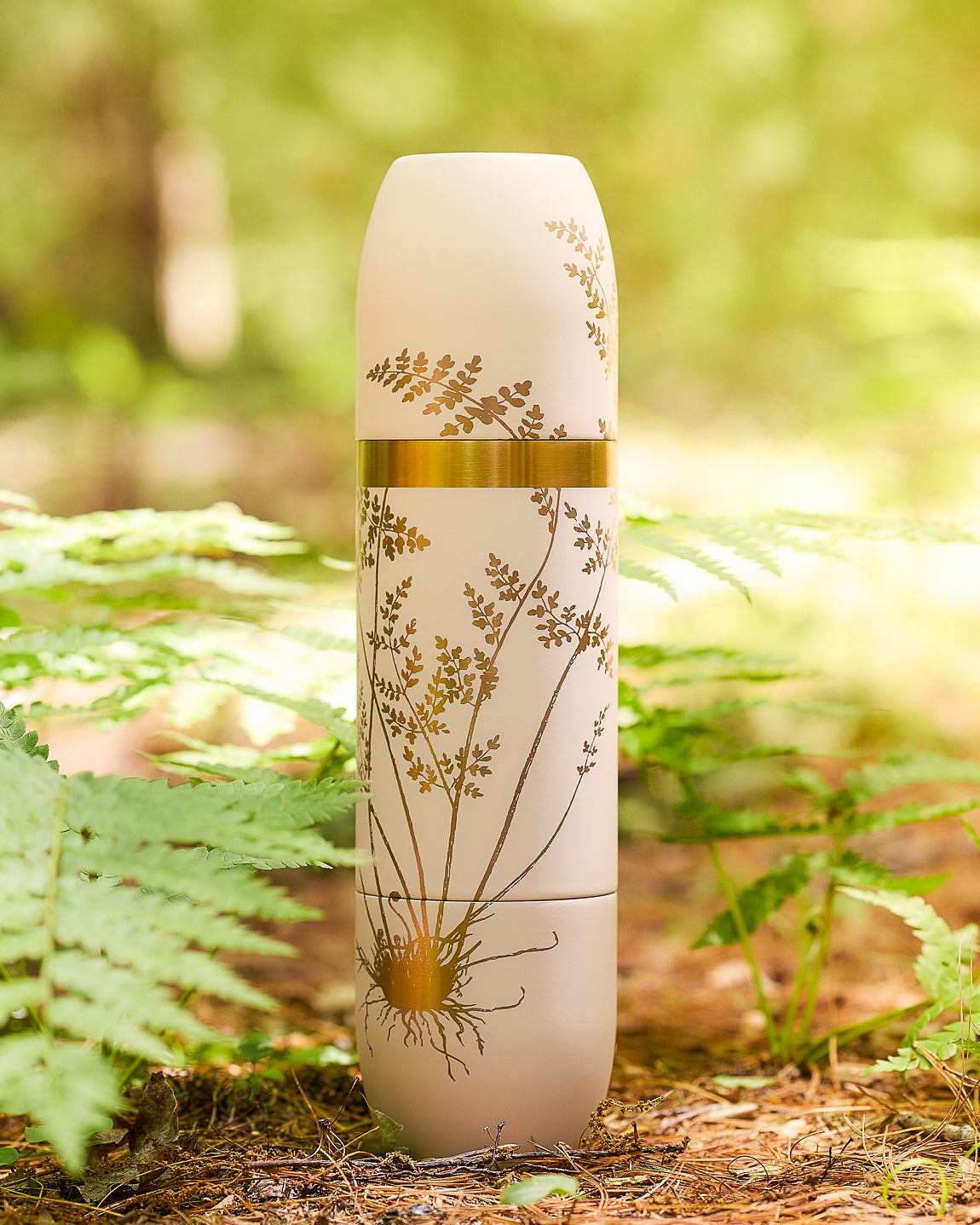 Caskata x High Camp wine Flask in Fairy Fern pattern engraved in gold on cream-colored flask on the forest floor amidst the ferns.