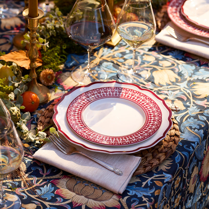 A table set with Caskata’s Stella Scalloped Crimson Dinner Plates, wine glasses, a fork on a napkin, and a floral tablecloth, adorned with greenery and candles for an heirloom-inspired display.
