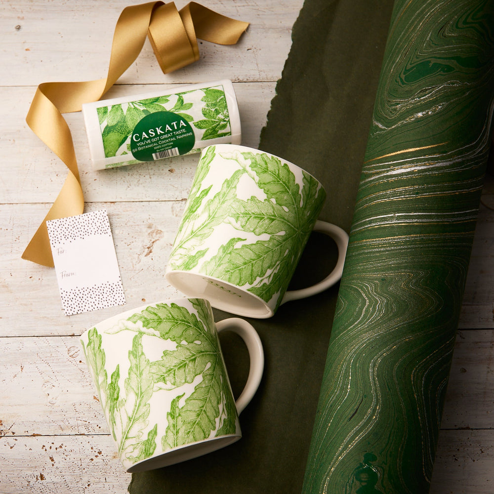 The Caskata Freya Coffee Talk Bundle, featuring two oversized mugs with a green botanical motif, green marbled wrapping paper, gold ribbon, a gift tag, and a branded box, is arranged on a white wooden surface.