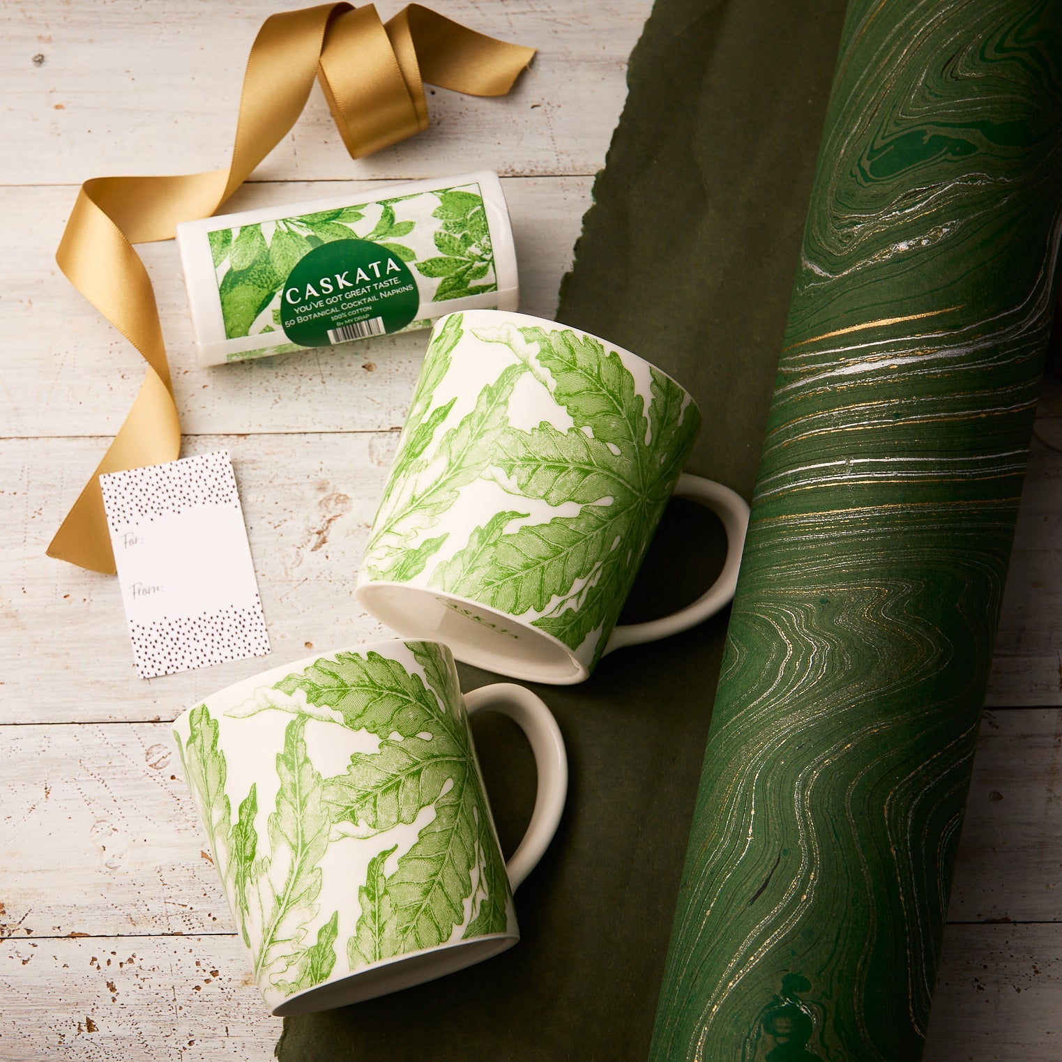 The Caskata Freya Coffee Talk Bundle, featuring two oversized mugs with a green botanical motif, green marbled wrapping paper, gold ribbon, a gift tag, and a branded box, is arranged on a white wooden surface.