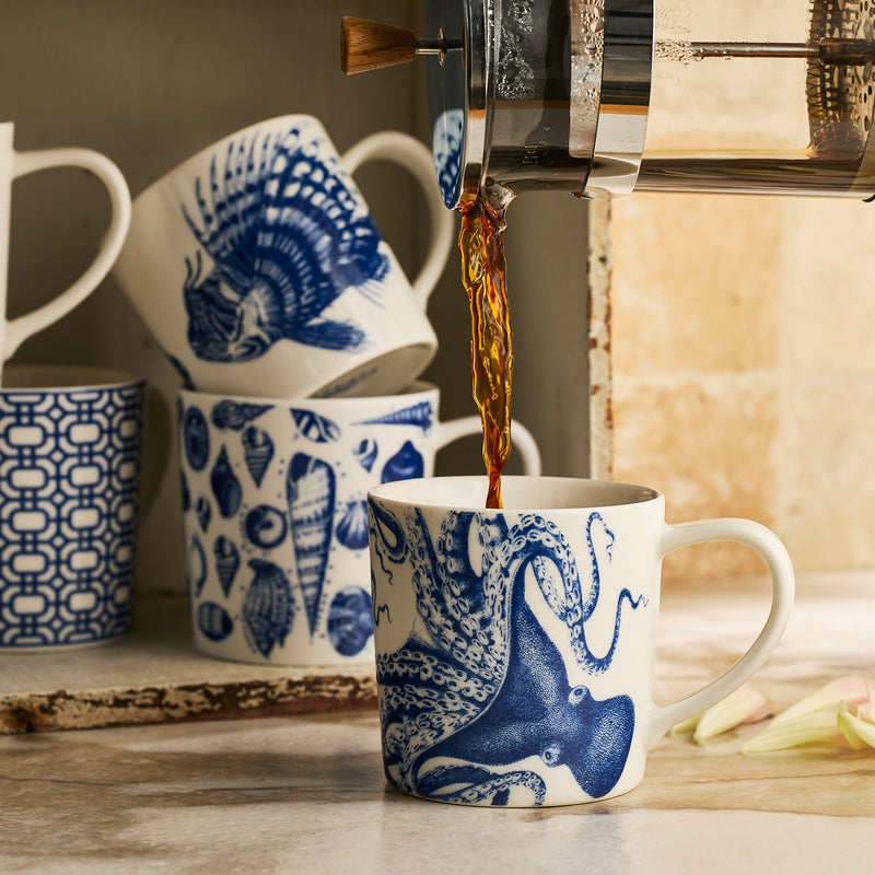 Coffee pours from a French press into the Lucy Mug by Caskata Artisanal Home, its striking blue marine design standing out among coastal drinkware and geometric patterned porcelain mugs.