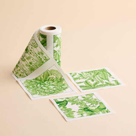 A roll of the MY DRAP Botanicals Verde Cocktail Napkin Roll, featuring green botanical designs, is partially unrolled with three square sections displayed flat on a beige surface—showcasing the fresh elegance typically found in floral cocktail napkin patterns.