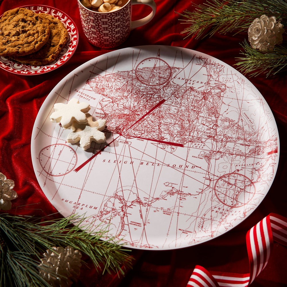 The Caskata White Holiday Hunt Large Round Tray holds two star-shaped cookies, set on a red tablecloth with more cookies, a mug, pine branches, and ribbon nearby.