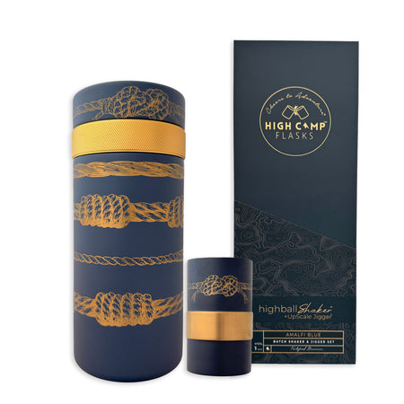 The Caskata x High Camp Sailors Knots Batch Cocktail Shaker, a cylindrical stainless steel flask with gold rope designs, is displayed beside its matching High Camp Flasks "Highball Shaker - Amalfi Blue" box.