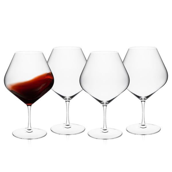 Four Caskata Library Red Wine Glasses, Set of 4, are arranged side by side; three are empty and one, featuring an oversized lead-free crystal bowl, is partially filled with red wine being gently swirled.