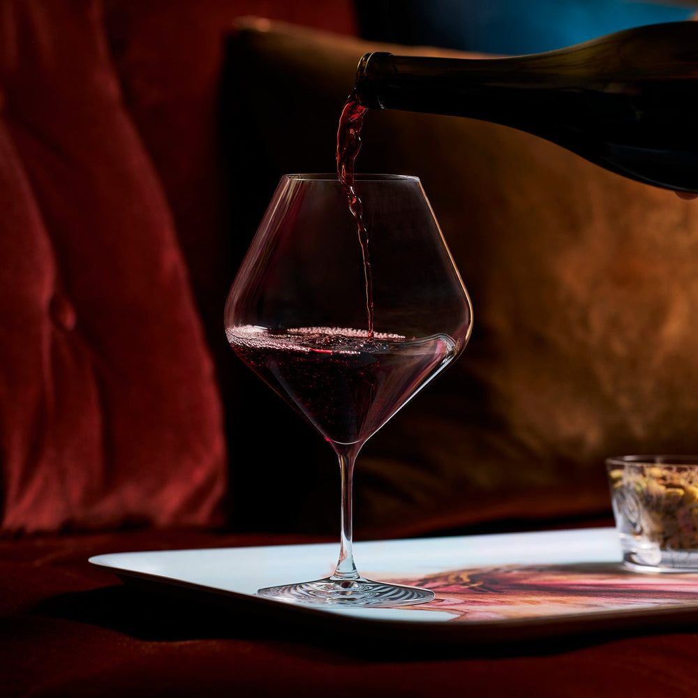 A bottle pours red wine into an oversized bowl of a Caskata Library Red Wine Glass (Set of 4) on a tray, with a small glass nearby and velvet cushions in the background.