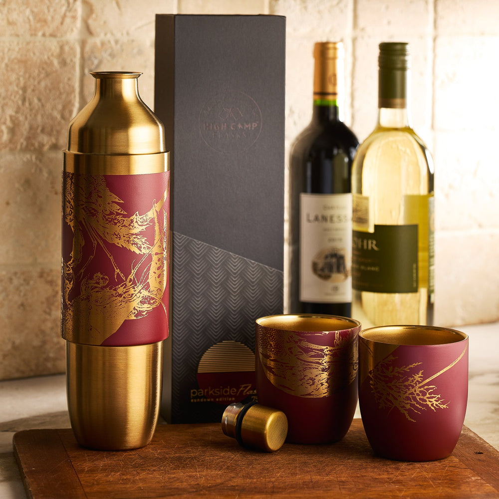 The Caskata x High Camp Red Lobster Wine Flask by Caskata is displayed on a kitchen counter, accompanied by two matching magnetized tumblers, a black gift box, and two bottles of wine.