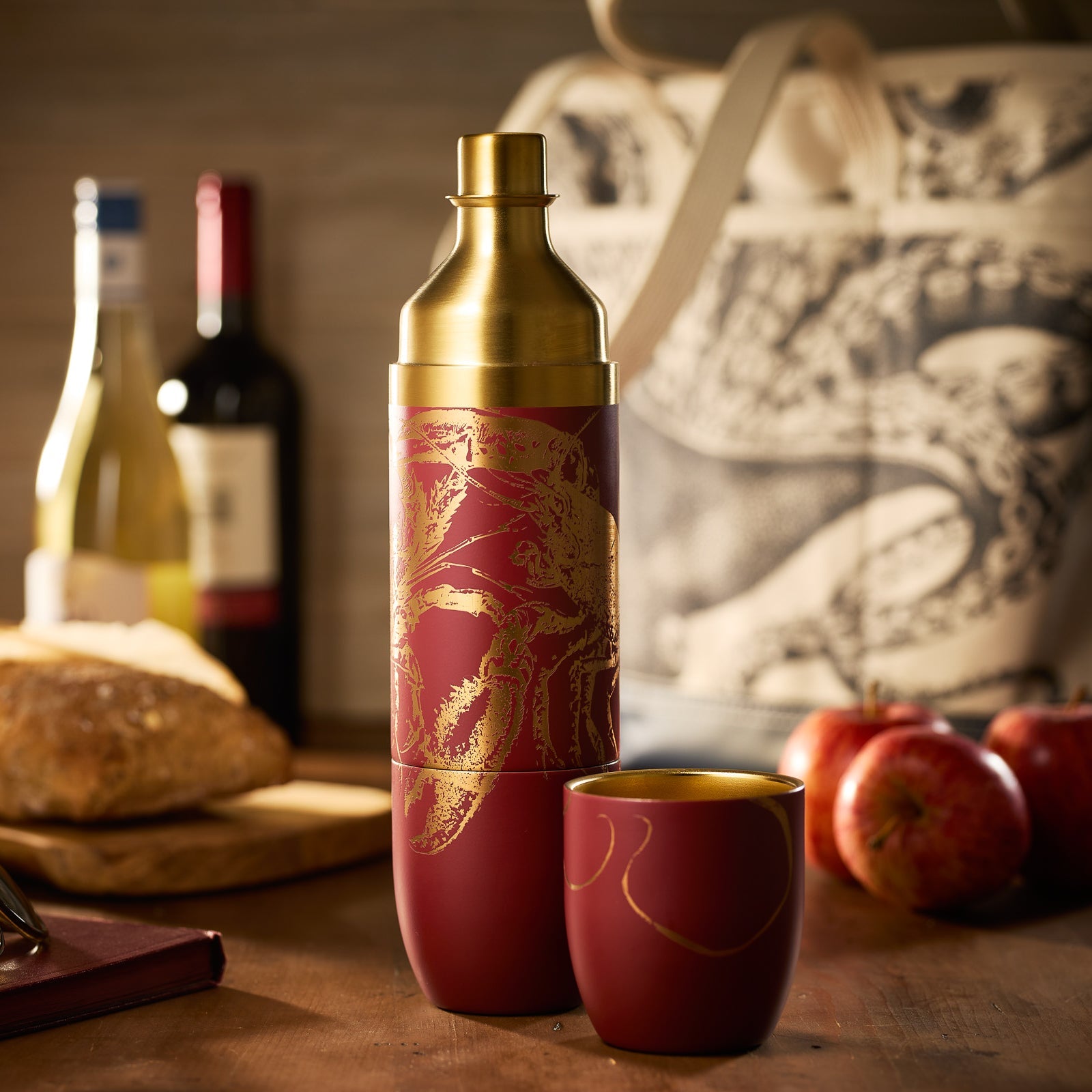 The Caskata X High Camp Flasks Red Lobster Flask stands upright in front of its gray felt carrying case with a brown label, showcasing its red stainless steel build and gold lobster design.