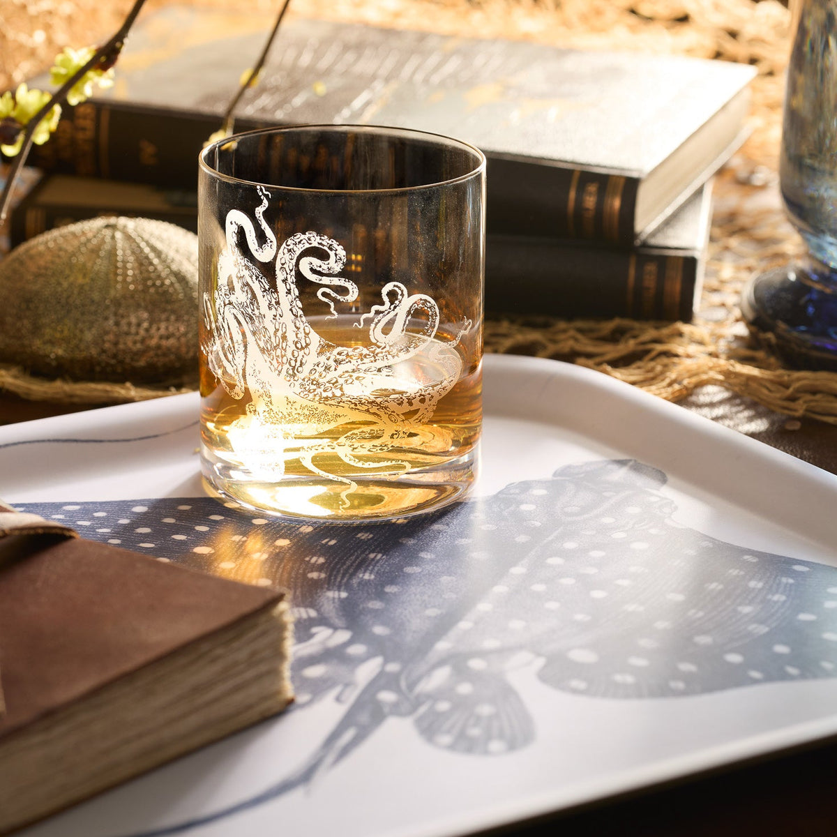 The Lucy Rocks Glass by Caskata Artisanal Home, featuring a sand-etched octopus design, sits partially filled on a fish-illustrated tray beside sea-inspired decor and books.