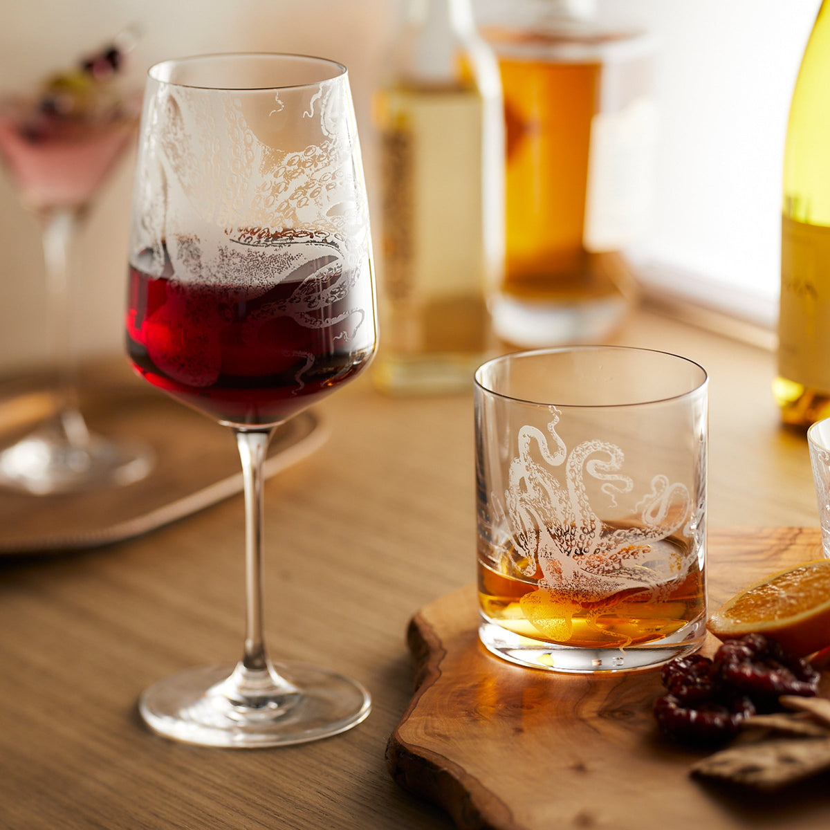A glass of red wine and a whiskey with ice rest elegantly on the wooden table, accompanied by bottles, oranges, and a small plate of food. The scene is framed by exquisite Lucy Red Wine Glasses from Caskata Artisanal Home that add an artistic flair to the display.