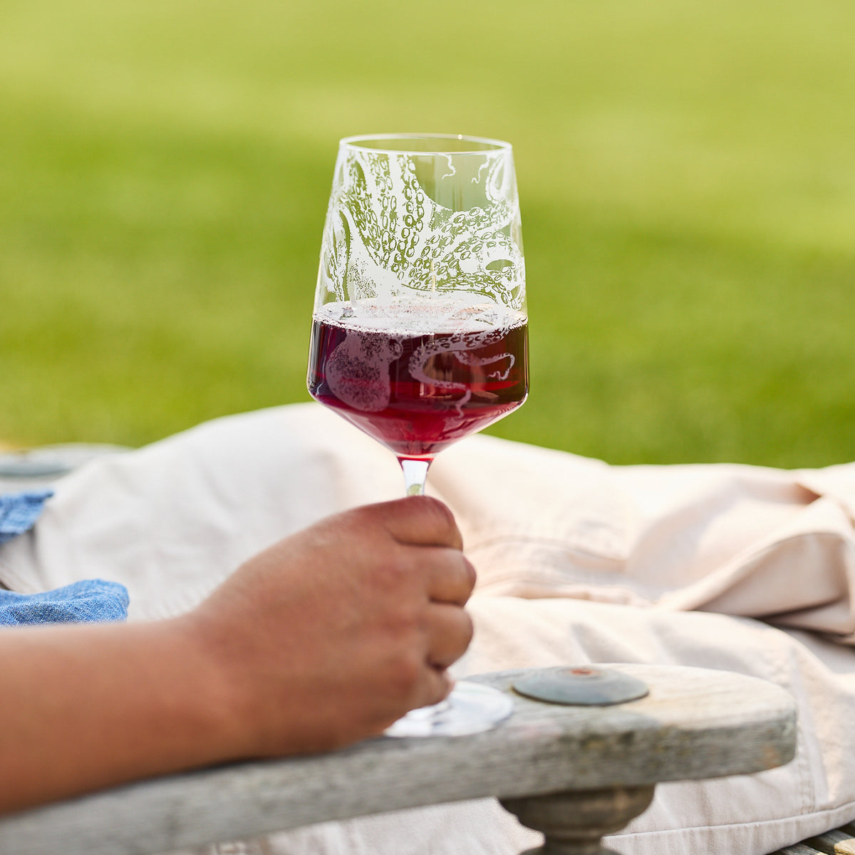 A person reclines outdoors on a wooden chair, holding a Caskata Artisanal Home Lucy Red Wine Glass, with green grass in the background.