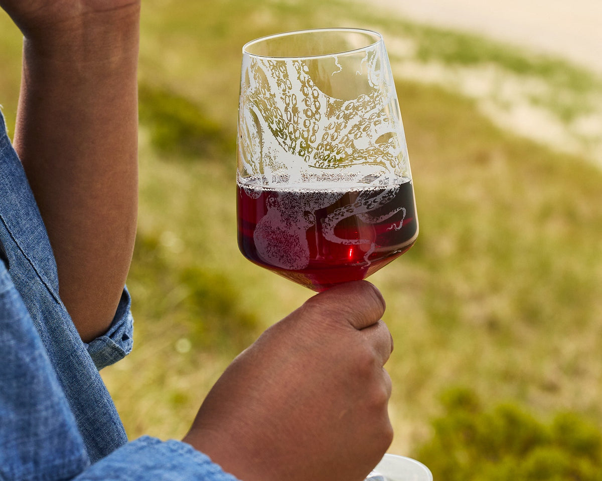 A person in a blue shirt holds a Caskata Artisanal Home Lucy Red Wine Glass outdoors, with grass and sand blurred in the background.