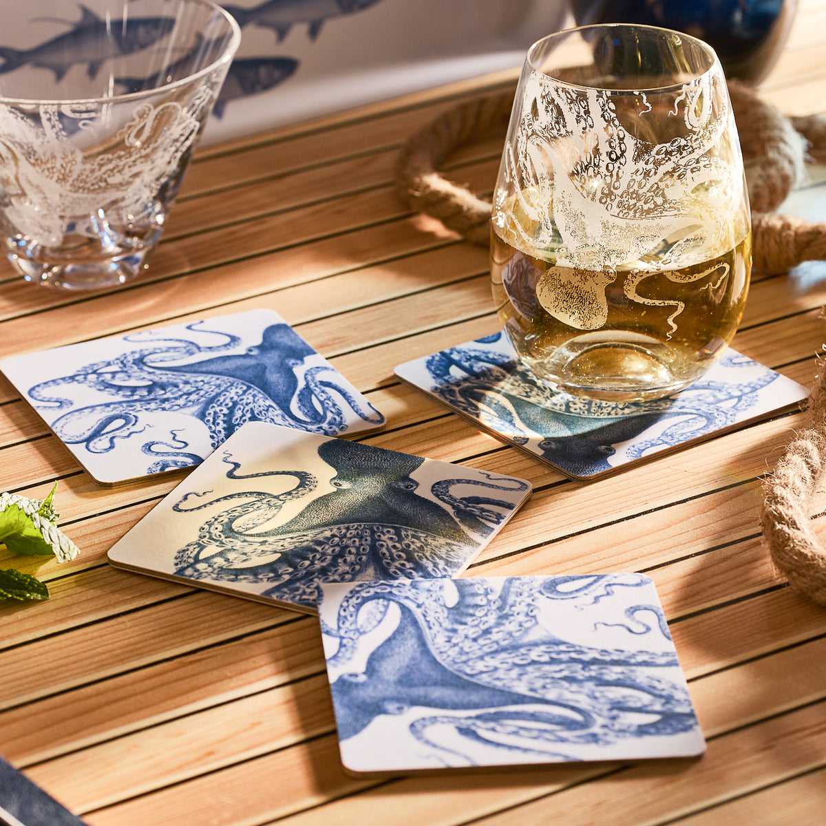 Caskata's Lucy Birchwood Coasters, a set of four with octopus designs, rest on a wooden table in sunlight, shadows dancing playfully; a nautical theme with rope and plants sets the scene.