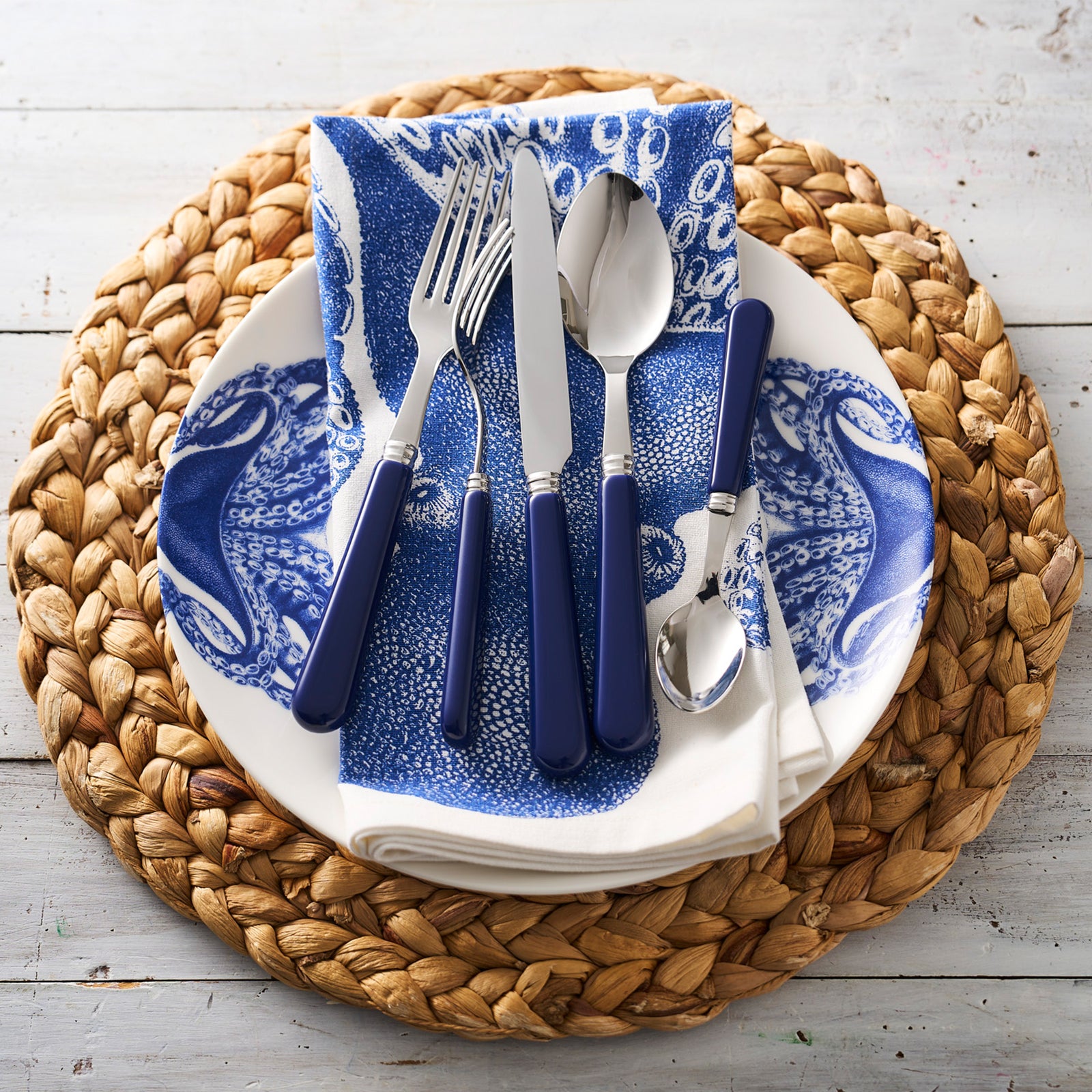 The Caskata Lucy Ultimate Set, a blue and white porcelain dish with an octopus design, holds a blue-patterned napkin and silverware on a woven placemat atop a light wood table.