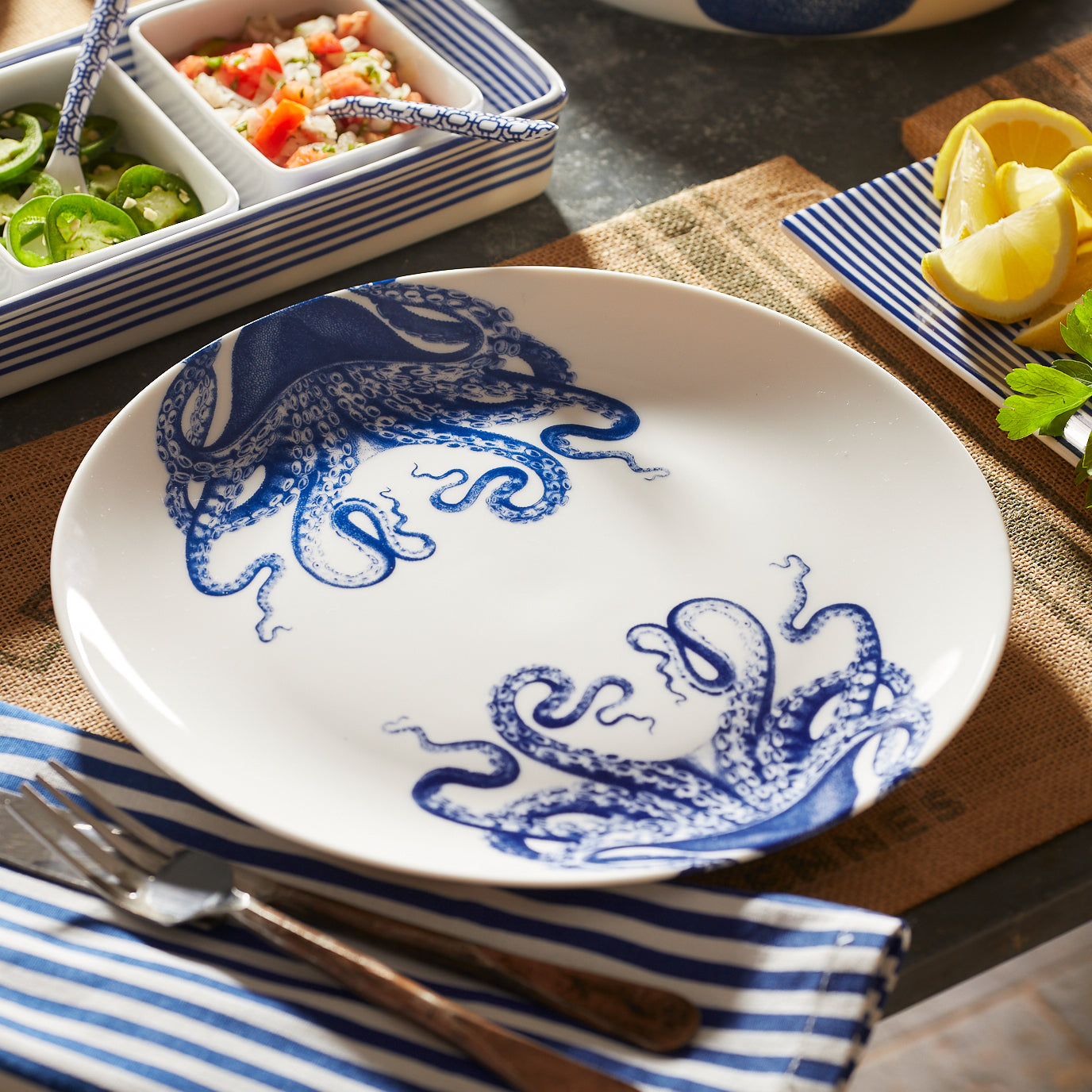 A Caskata "Lucy Table for 8" white plate, decorated with blue octopus illustrations, is displayed on a table with striped napkins, cutlery, lemon slices, and bowls of vegetables.