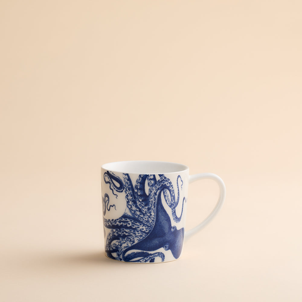 The Lucy Mug by Caskata Artisanal Home features a charming blue octopus illustration on white porcelain, displayed against a simple beige surface and background—perfect for bringing a coastal vibe to your morning routine.