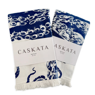 Two folded Caskata Lucy table runners, white cotton with blue octopus tentacle designs, each wrapped in branded labels—perfect for coastal table decor.