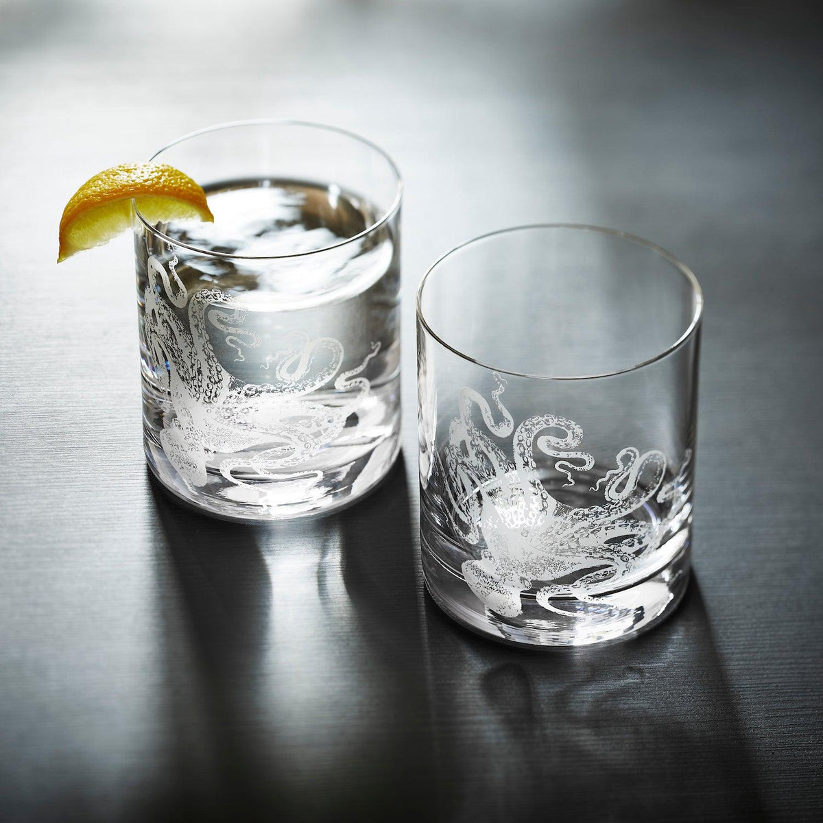 Two Caskata Artisanal Home Lucy Rocks Glasses with octopus engravings, one filled with water and garnished with a lemon wedge, the other empty, placed on a dark surface. The intricate design showcases the artistry of the sand-etching technique used to create this exquisite octopus glassware.